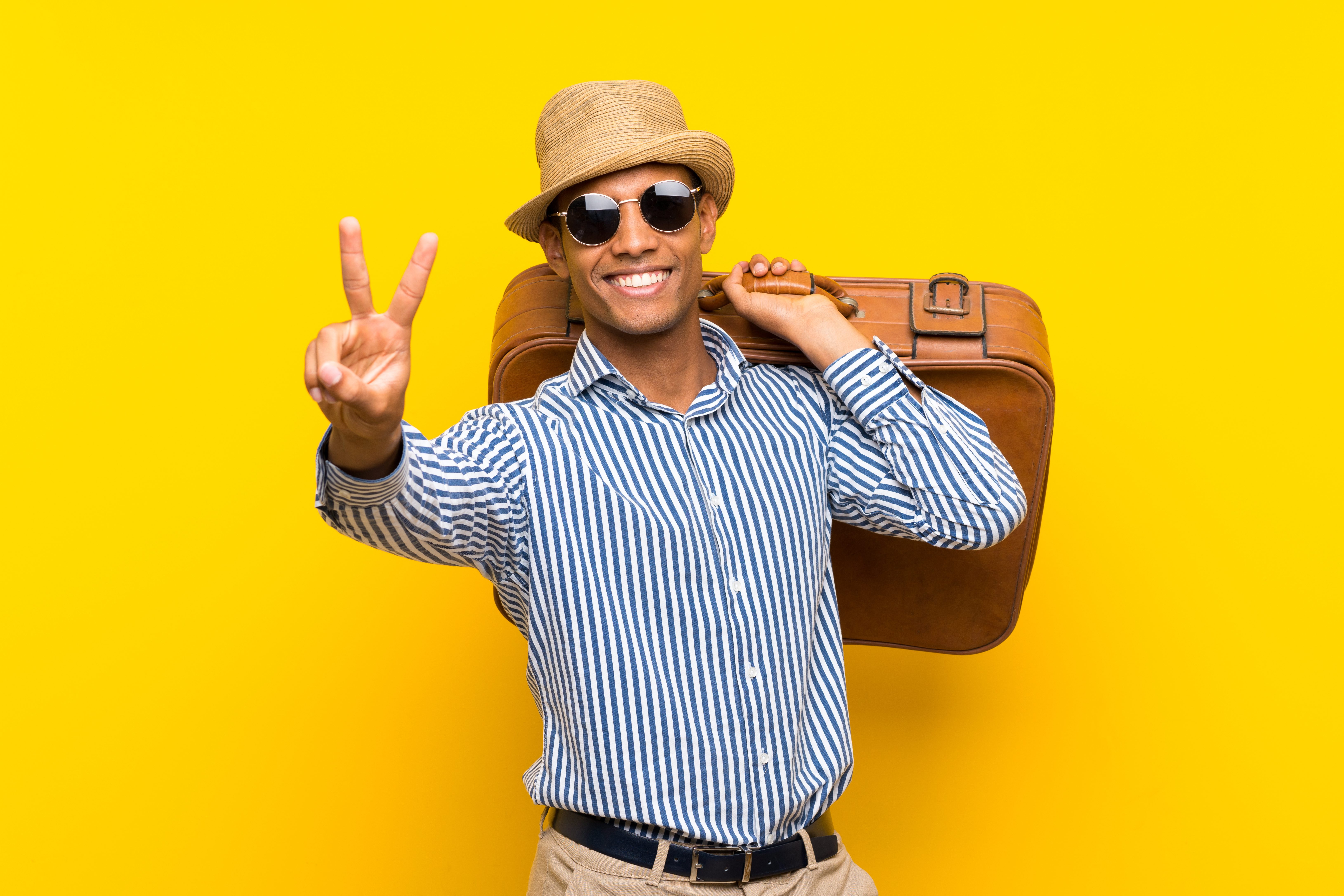 Brunette man holding a vintage briefcase over isolated yellow background smiling and showing victory sign