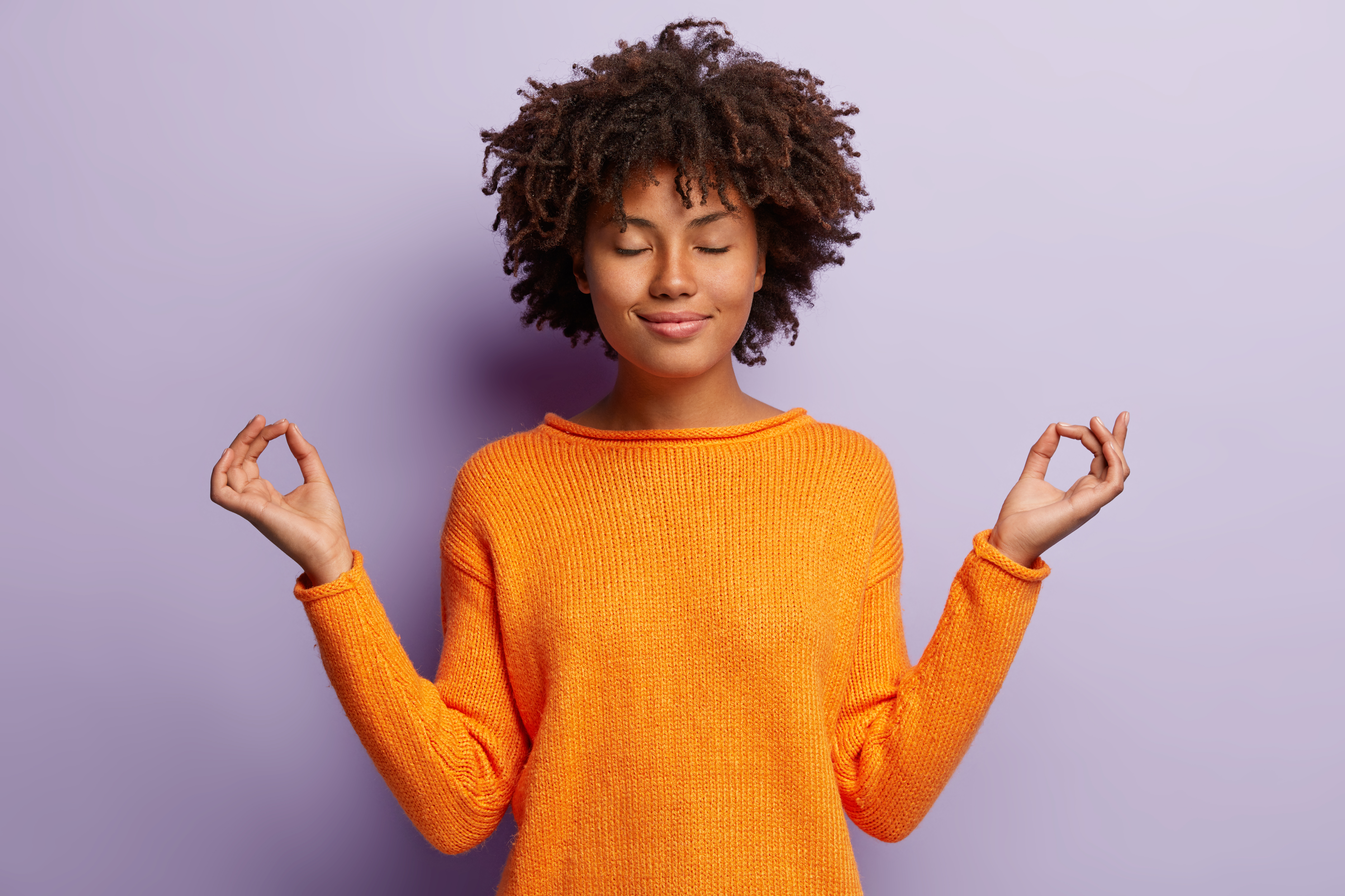 Pleasant looking calm woman meditates indoor, holds hands in mudra gesture, has charming smile, closed eyes, wears orange clothes, models over purple background. Hand gesture. Meditation concept