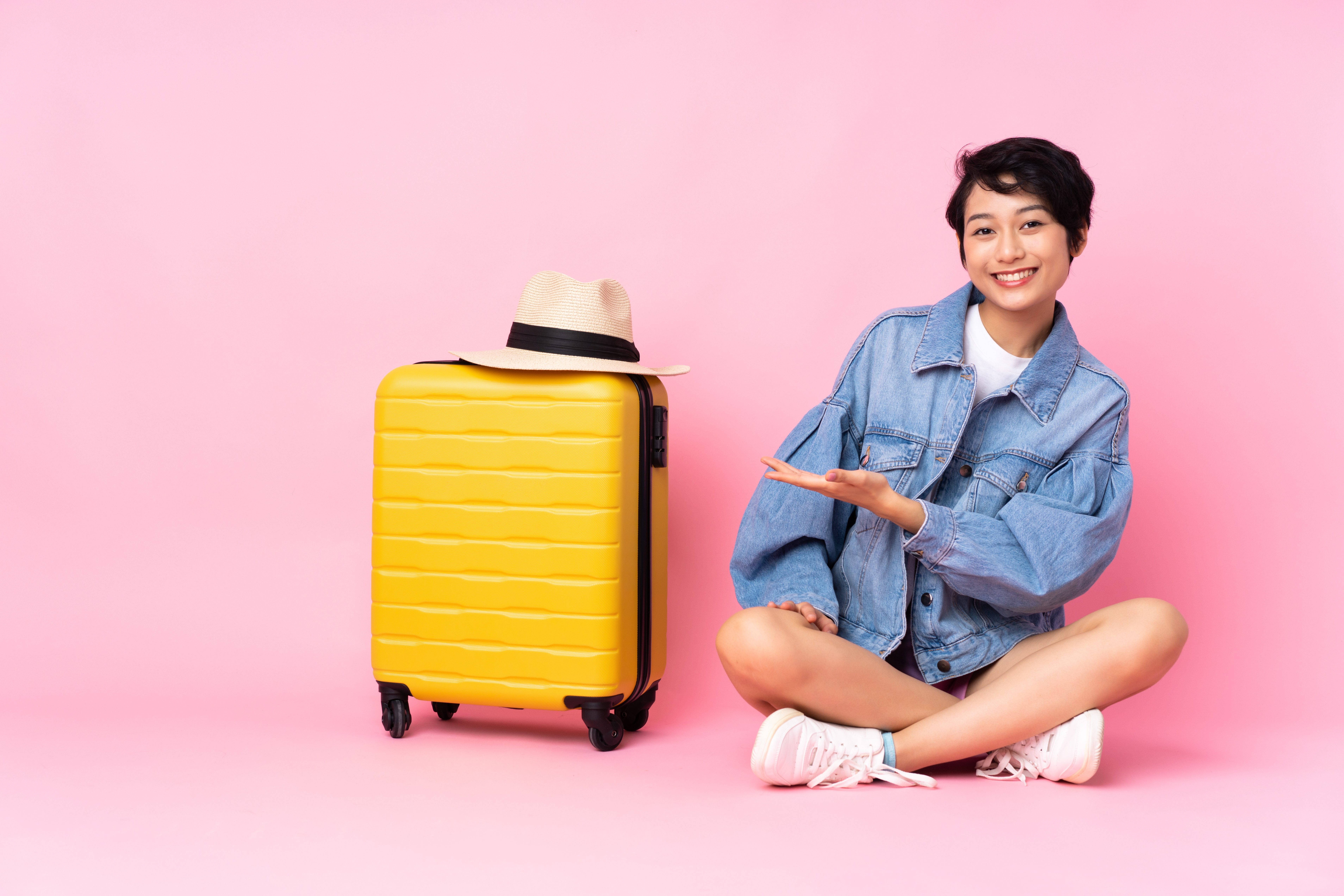 Young traveler Vietnamese woman with suitcase sitting on the floor over isolated pink background presenting an idea while looking smiling towards
